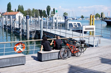 Ein Radlerpärchen sitzt auf einer Bank und genießt den Blick auf's Wasser; im Hintergrund ist das Zollhaus zu erkennen, das als ältestes Lagerhaus in Romanshorn gilt.