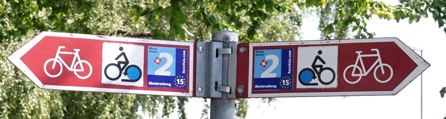 Roter Doppel-Wegweiser entlang des Radwegs mit stilisiertem Fahrrad und dem Logo der Rhein-Route.